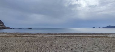 Bulutlu gökyüzünün altında sakin bir sahil manzarası, yumuşak dalgalar ve kumlu bir kıyı, uzak tepeler tarafından kuşatılmış