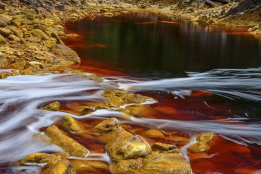 Foaming water rushes over the colorful, iron-laden rocks in the Rio Tinto river clipart
