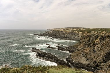 Portekiz, Cabo Sardao 'daki görkemli kayalıklar, engebeli kaya oluşumları ve dalgalar kıyıya çarpıyor. Parçalı bulutlu bir gökyüzü dramatik kıyı manzarasını güçlendirir.