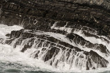Portekiz 'deki Cabo Sardao' da dalgaların sert kayalara çarpmasının yakın görüntüsü. Çalkantılı deniz ve kaya oluşumları dinamik ve güçlü bir sahne yaratır..