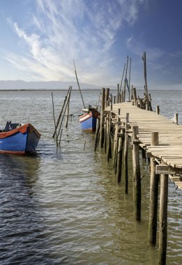 Yıpranmış ahşap bir iskele ve küçük bir kulübe yakınlarda demir atmış bir tekneyle sakin bir nehre uzanıyor..