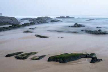 Portekiz 'deki Amoreira Sahili' nde sakin bir sahne. Sis kaplı kayalar ve kumlu kıyıya hafifçe vuran yumuşak dalgalar. Sakin atmosfer sisli ve sakin denizle güçlenir..