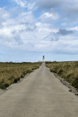 Menorca 'daki tarihi Faro de Cavalleria deniz fenerine giden uzun, düz bir yol parçalı bulutlu bir gökyüzüne doğru ilerliyor. Seyahat ve manzara temaları için ideal.