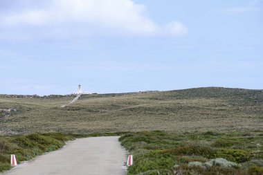 Menorca 'daki tarihi Faro de Cavalleria deniz fenerine giden uzun, düz bir yol parçalı bulutlu bir gökyüzüne doğru ilerliyor. Seyahat ve manzara temaları için ideal.