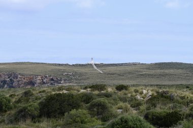 Menorca 'daki tarihi Faro de Cavalleria deniz fenerine giden uzun, düz bir yol parçalı bulutlu bir gökyüzüne doğru ilerliyor. Seyahat ve manzara temaları için ideal.