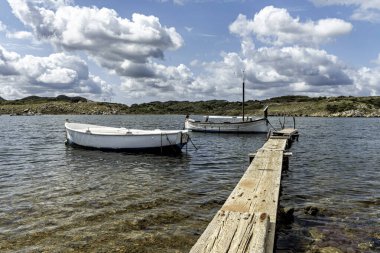 Sanitja Limanı, Menorca 'nın berrak sularına yanaşan teknelere giden ahşap iskele. Parçalı bulutlu bir gökyüzünün altında kayalık kıyı şeridi ve yemyeşil manzarası.