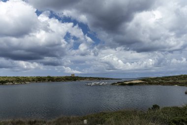 Sanitja Limanı, Menorca 'da sakin sularda demirli teknelerin olduğu pitoresk bir manzara. Manzara kayalık kıyılar, yemyeşil yemyeşil ve parçalı bulutlu bir gökyüzünün altında eski bir taş yapıya sahiptir..