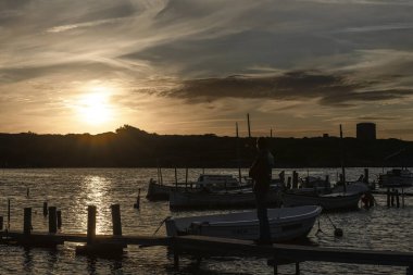 Menorca 'nın Sanitja Limanı' nın sularında, iskeleye demirlemiş geleneksel teknelerle baş döndürücü bir günbatımı. Gökyüzü sıcacık renklerle boyanır. Huzurlu ve güzel bir sahil manzarası yaratır..