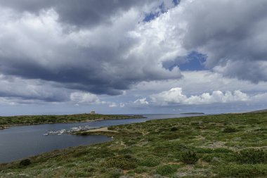 Sanitja Limanı, Menorca 'da sakin sularda demirli teknelerin olduğu pitoresk bir manzara. Manzara kayalık kıyılar, yemyeşil yemyeşil ve parçalı bulutlu bir gökyüzünün altında eski bir taş yapıya sahiptir..