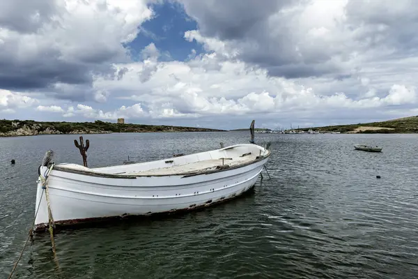 stock image Wooden fishing boats moored at the pier in Port of Sanitja, Menorca. The tranquil water and lush, green landscape under a partly cloudy sky create a serene coastal scene.