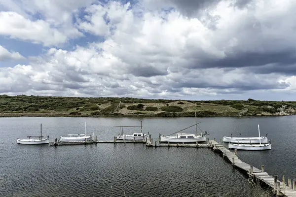 stock image Wooden pier leading to boats docked in the clear waters of Port of Sanitja, Menorca. Scenic view of the rocky shoreline and lush greenery under a partly cloudy sky.