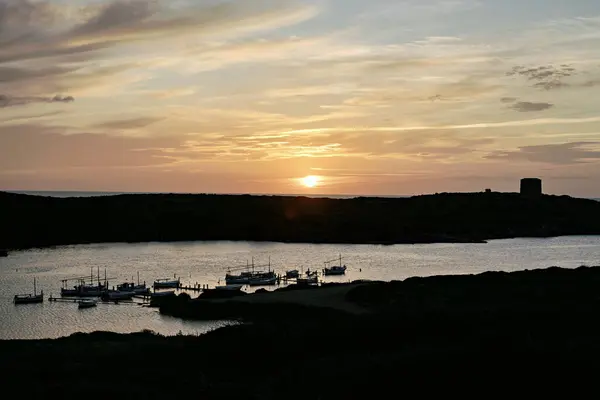 stock image A stunning sunset over the waters of Port of Sanitja, Menorca, with traditional boats moored at the pier. The sky is painted with warm hues, creating a serene and picturesque coastal scene.