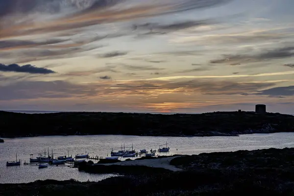 stock image A stunning sunset over the waters of Port of Sanitja, Menorca, with traditional boats moored at the pier. The sky is painted with warm hues, creating a serene and picturesque coastal scene.