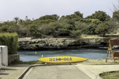 Sarı sörf tahtası, Menorca 'daki Cala Blanca Plajı' nda suyun kenarında dinlenen Hola Ola yazısı. Kayalık kıyı şeridinin manzarası ve yemyeşil.