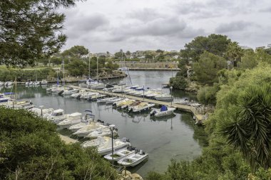 Menorca 'daki Cala Busquets Marina' ya kenetlenmiş çeşitli teknelerin manzarası. Arkaplanda yemyeşil ve konut binaları bulutlu bir gökyüzünün altında.