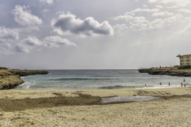 Ziyaretçiler, Menorca 'daki Cala en Bosch plajında güneşli bir günün tadını çıkarıyorlar. Kumlu sahil, sakin sular ve parçalı bulutlu gökyüzü dinlenmek ve dinlenmek için ideal bir ortam yaratır..