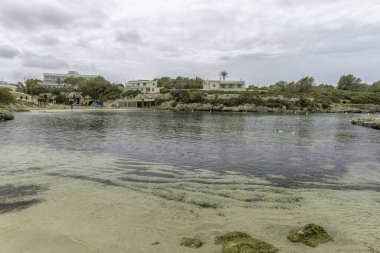 Menorca 'daki Cala Santandria' nın berrak, sakin suları, kayalık kıyıları ve yemyeşil arazisiyle huzurlu manzarası. Sarı şamandıralar bulutlu gökyüzünün altındaki suyu işaret ediyor..