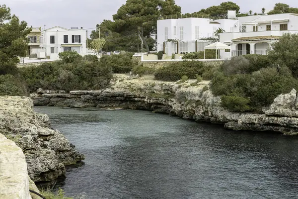 Tarihi bir kule ve modern binaların yer aldığı Cala Busquets, Menorca 'daki sahil sahnesi. Kayalık kayalıklar ve sakin sular bulutlu bir gökyüzünün altında.