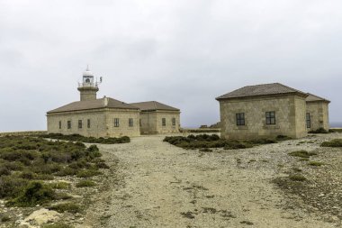 Menorca 'daki tarihi Punta Nati Deniz feneri kompleksi bulutlu bir gökyüzü altında taş binalar ve deniz feneri barındırıyor. Sakin ve engebeli bir sahil manzarası.