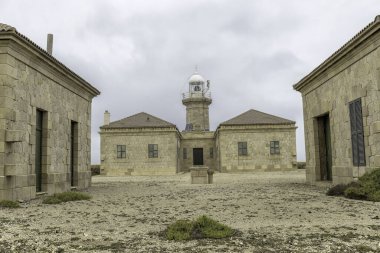 Menorca 'daki tarihi Punta Nati Deniz feneri kompleksi bulutlu bir gökyüzü altında taş binalar ve deniz feneri barındırıyor. Sakin ve engebeli bir sahil manzarası.