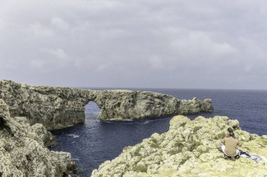 Menorca 'daki Pont de Gil' in göz kamaştırıcı manzarası, bulutlu bir gökyüzünün altında derin mavi denize uzanan doğal bir kaya kemeri. Muhteşem bir kıyı düzeni..