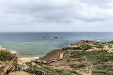 Menorca 'daki Cala Pilar' ın sakin bir sahnesinde altından kumlu tenha bir sahil ve bulutlu bir gökyüzünün altında engebeli kayalık bir tepe..