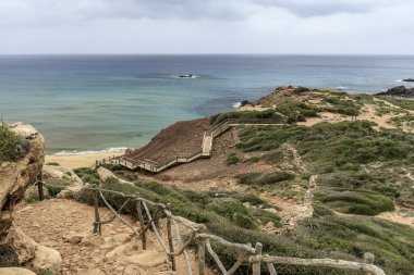 Menorca 'daki Cala Pilar' ın sakin bir sahnesinde altından kumlu tenha bir sahil ve bulutlu bir gökyüzünün altında engebeli kayalık bir tepe..