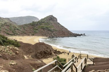 Menorca 'daki Cala Pilar' ın sakin bir sahnesinde altından kumlu tenha bir sahil ve bulutlu bir gökyüzünün altında engebeli kayalık bir tepe..