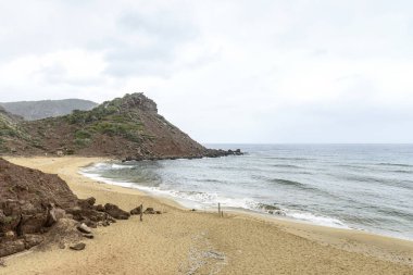 Menorca 'daki Cala Pilar' ın sakin bir sahnesinde altından kumlu tenha bir sahil ve bulutlu bir gökyüzünün altında engebeli kayalık bir tepe..