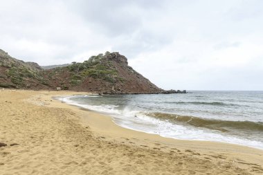 Menorca 'daki Cala Pilar' ın sakin bir sahnesinde altından kumlu tenha bir sahil ve bulutlu bir gökyüzünün altında engebeli kayalık bir tepe..
