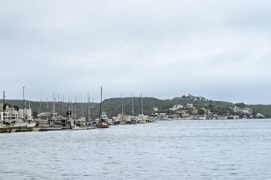 Menorca 'daki Mahon limanının sakin manzarasında, bulutlu bir gökyüzü altında sakin sular, rıhtımlı tekneler ve çevreleyen tepeler.