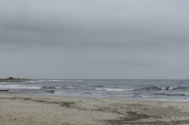 Menorca 'daki Punta Prima Sahili' nin huzurlu manzarası kumlu bir sahili, yumuşak dalgaları ve bulutlu bir gökyüzü altında uzak bir yelkenliyi gösteriyor..