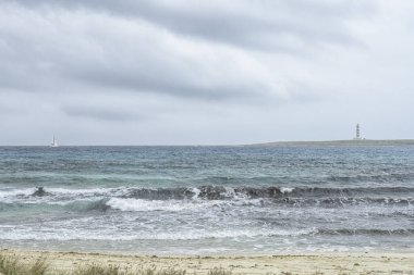 Menorca 'daki Punta Prima Plajı' nda sakin bir sahne, kumlu kıyılar, hafif dalgalar ve bulutlu bir gökyüzünün altında bir deniz feneri manzarası..