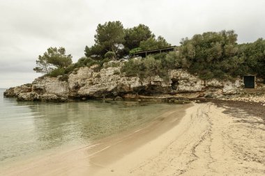 Menorca 'daki Cala Benissafe' in sakin bir manzarası, sakin bir koy, kumlu bir sahil, kayalık kayalıklar ve küçük beyaz bir ev. Bulutlu gökyüzü huzur dolu bir atmosfer ekler..