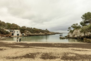 Menorca 'daki Cala Benissafe' in sakin bir manzarası, sakin bir koy, kumlu bir sahil, kayalık kayalıklar ve küçük beyaz bir ev. Bulutlu gökyüzü huzur dolu bir atmosfer ekler..