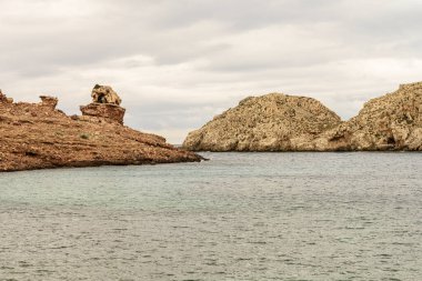 Cala Morell, Menorca 'nın kendine özgü kayalık oluşumları bulutlu bir gökyüzünün altında. Engebeli arazi sakin Akdeniz 'le buluşuyor..
