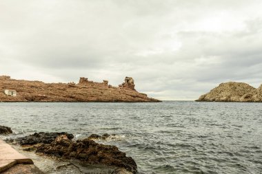Cala Morell, Menorca 'nın kendine özgü kayalık oluşumları bulutlu bir gökyüzünün altında. Engebeli arazi sakin Akdeniz 'le buluşuyor..