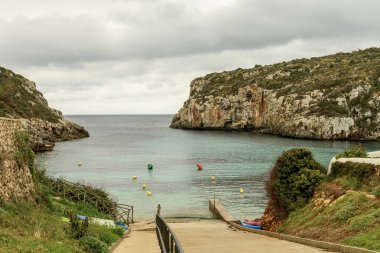 Menorca 'daki Cala Es Canutells' in sakin manzarasında bulutlu bir gökyüzü altında turkuaz sular, kayalık uçurumlar ve renkli şamandıralar yer alıyor. Huzurlu bir Akdeniz Koyu.