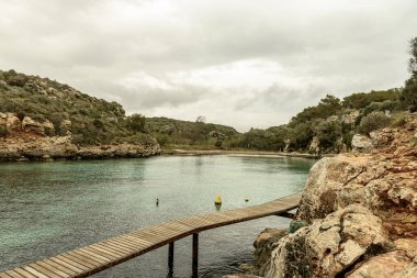 Menorca 'daki Cala Es Canutells' in sakin manzarasında bulutlu bir gökyüzü altında turkuaz sular, kayalık uçurumlar ve renkli şamandıralar yer alıyor. Huzurlu bir Akdeniz Koyu.