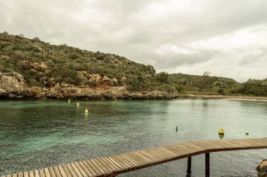 Menorca 'daki Cala Es Canutells' in sakin manzarasında bulutlu bir gökyüzü altında turkuaz sular, kayalık uçurumlar ve renkli şamandıralar yer alıyor. Huzurlu bir Akdeniz Koyu.