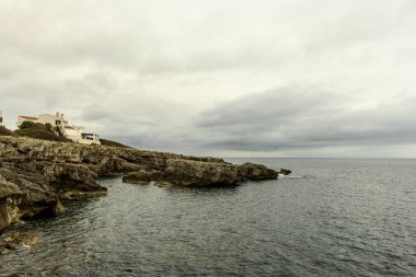 Binibeca, Menorca 'da huzurlu bir deniz manzarası, engebeli kayalıkların yanında bir yelkenli ve bulutlu bir gökyüzünün altında sakin sular..