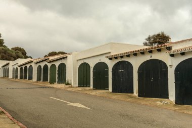 Binibeca, Menorca 'da, bulutlu bir gökyüzünün altında kemerli kapılı geleneksel kayıkhaneler. Huzurlu ve tarihi mimari, kıyı köyüne cazibe katıyor..