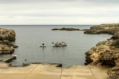 Binibeca, Menorca 'daki balıkçı limanının huzurlu manzarasında sakin sularda yüzen küçük tekneler, kayalık kıyı şeridi ve bulutlu bir gökyüzü var..