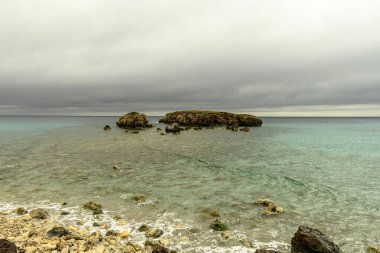 Menorca 'daki Sant Adeodat Sahili' nin huzurlu manzarasında kayalık bir kıyı şeridi, berrak turkuaz sular ve bulutlu bir gökyüzü yer alıyor. Kıyı boyunca uzanan ahşap bir patika..