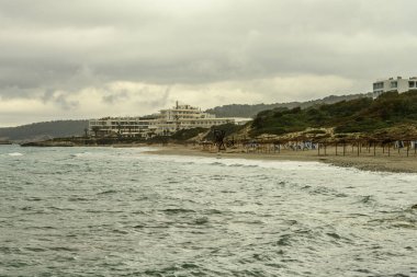 Menorca 'daki Santo Tomas Sahili' nin dramatik manzarası, bir çakıl taşı kıyı şeridi, turkuaz sular ve yaklaşan fırtına bulutları. Sahile yakın binalar ve tahta kaldırımlar eklendi..