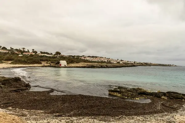 stock image A rustic beach hut stands by the rocky shoreline of Cala Binibeca, Menorca, under a cloudy sky.