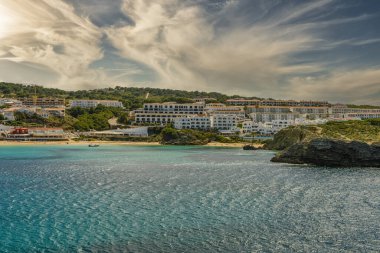 Menorca 'daki Arenal d' en Castell 'in çarpıcı manzarası, turkuvaz suları, kumlu bir plaj ve yemyeşil bir zemin ve dramatik bir gökyüzü karşısında beyaz Akdeniz tarzı binalar..