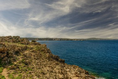 Menorca 'daki Arenal d' en Castell 'in çarpıcı manzarası, turkuvaz suları, kumlu bir plaj ve yemyeşil bir zemin ve dramatik bir gökyüzü karşısında beyaz Akdeniz tarzı binalar..