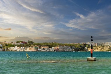 Menorca 'daki Cala Escull limanının manzarası, demirlemiş tekneler, berrak mavi sular ve parçalı bulutlu bir gökyüzünün altındaki kayalık bir kıyı şeridi..