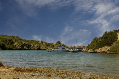 Menorca 'daki Cala Escull limanının manzarası, demirlemiş tekneler, berrak mavi sular ve parçalı bulutlu bir gökyüzünün altındaki kayalık bir kıyı şeridi..